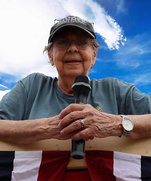 Sue Dyer on a podium holding a microphone at the Hamner Guideboat Race.